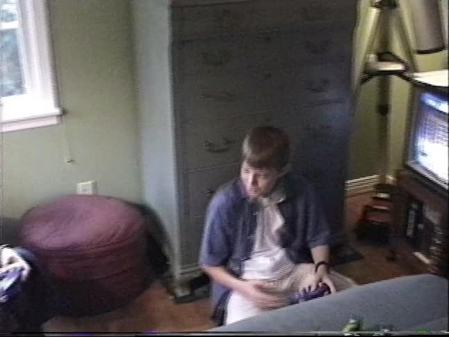 A vintage home video shows two young boys playing video games in a bedroom, with one pulling his shirt over his head in a funny moment