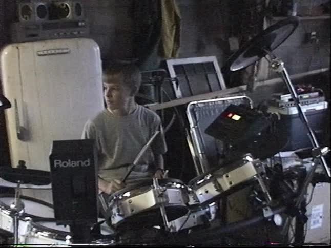 Vintage home video of a young boy smiling while practicing on his electronic drum set in a messy garage