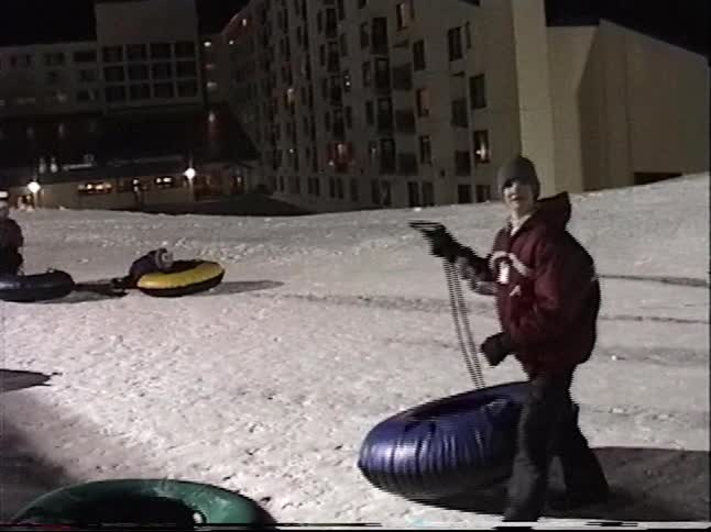 Vintage home movie captures a group of people preparing to go snow tubing at night on a snowy hill