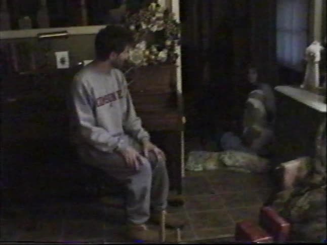 A man sits patiently in a dimly lit living room while a dog walks by in vintage home video footage