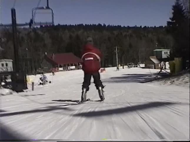 A point-of-view shot follows a skier in a red Alabama jacket down a gentle slope at a sunny ski resort