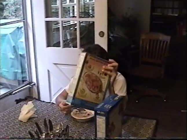 A boy hides behind a cereal box while eating breakfast at the dining table in a funny home video