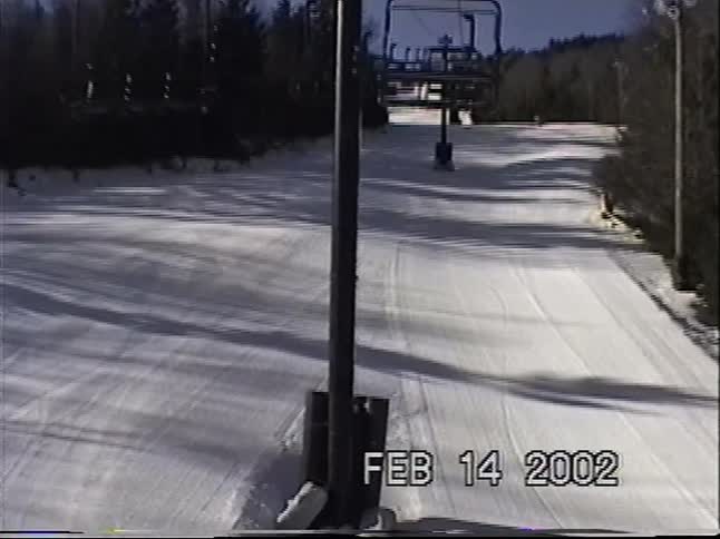A person wearing winter gear and sunglasses rides a ski lift up a sunny mountain in this vintage 2002 footage