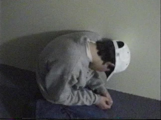 A grainy home video captures a young boy walking and jumping playfully down a long, dark hallway where his friend is found hiding in a corner