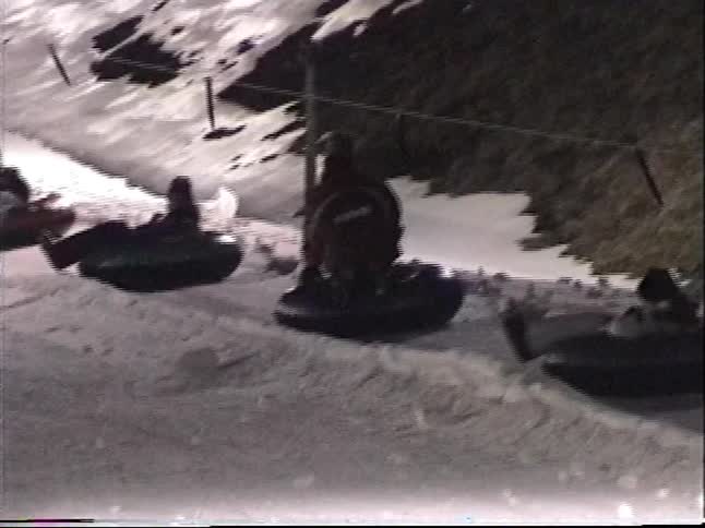 Vintage footage shows a line of people being towed up a brightly lit snow tubing course at a winter resort at night