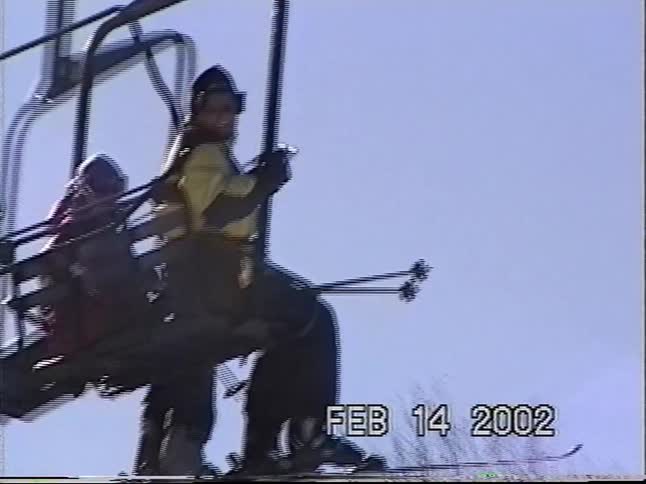 A father and son ride a chairlift up the snowy mountain on a sunny winter day, captured on vintage VHS footage from 2002