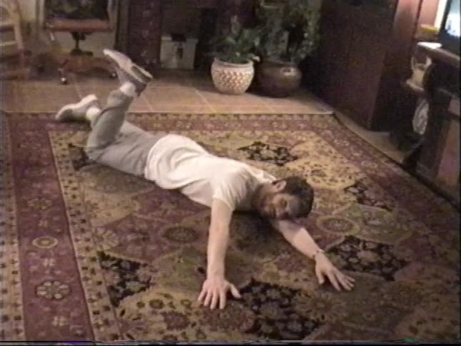 A silly man playfully pretends to swim across a large, patterned rug in his living room in this vintage home video