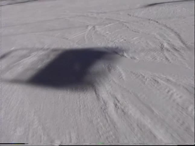 A vintage first-person perspective of skiing down a sunny mountain slope, passing a resting snowboarder before meeting a smiling young skier
