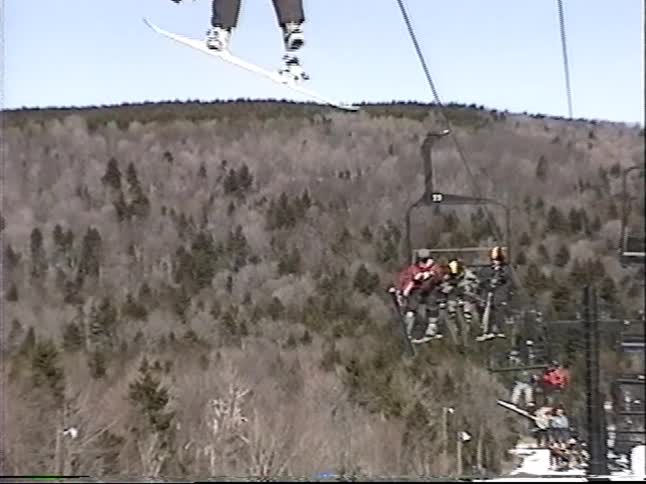 Point of view from a ski lift following chairs with skiers and snowboarders up a snowy mountain on a bright, sunny day