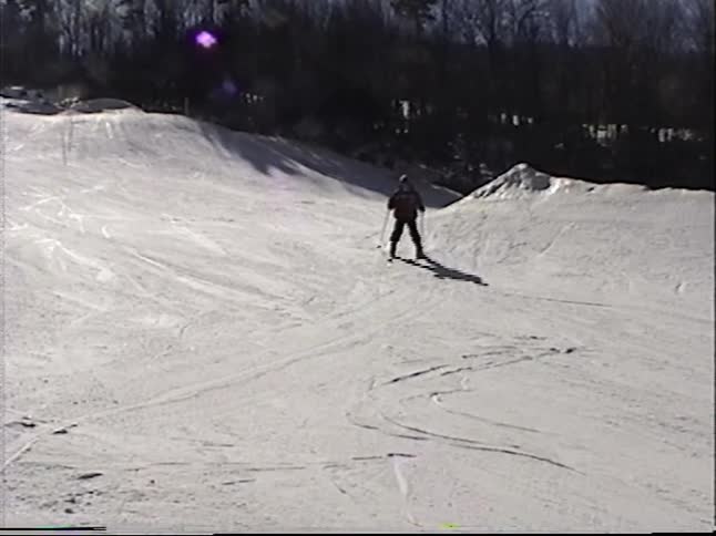 Vintage home video captures people skiing and snowboarding down a sunny mountain slope under a chairlift