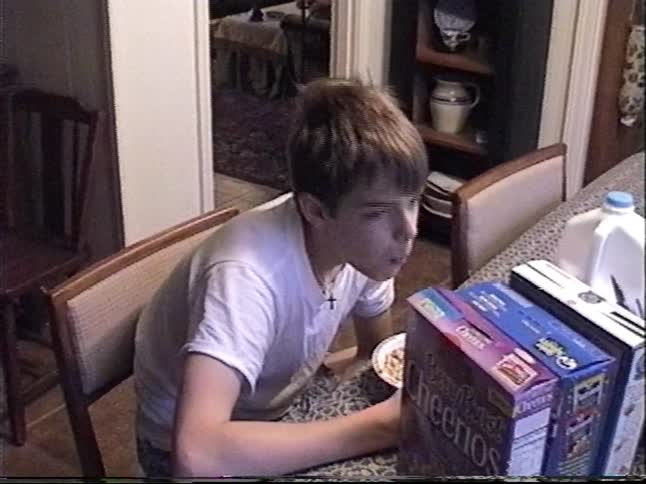 Vintage VHS footage shows a young boy eating breakfast and reading the cereal box at the dining table