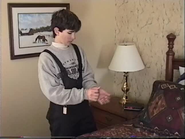 A vintage home video shows a teenage boy in snow bibs walking through a home and into his bedroom before sitting down on the bed