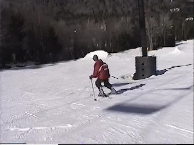 An older man in a red jacket enjoys a sunny day of skiing down a groomed mountain slope, filmed from a following skier's perspective