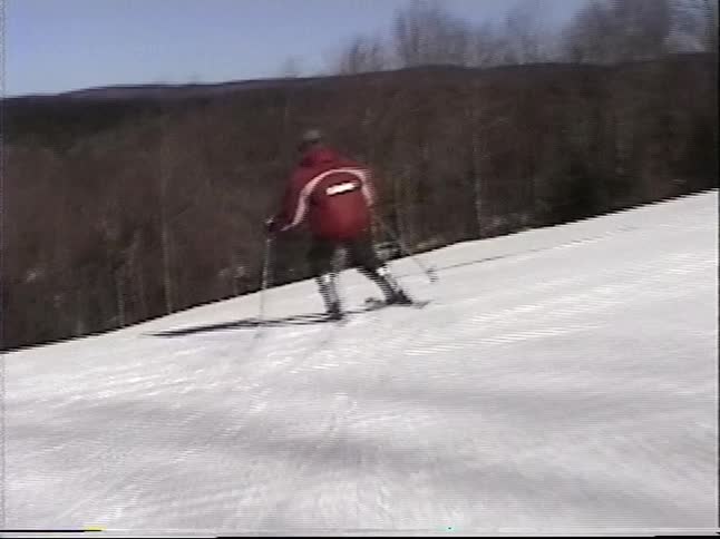 Point-of-view footage follows a skier in a red jacket as they ski down a groomed slope on a sunny winter day