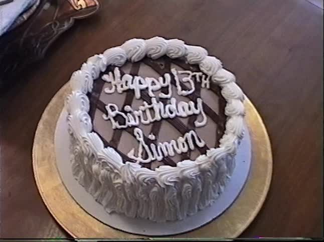 A close-up home video shot of a 'Happy 13th Birthday Simon' cake on a dining room table