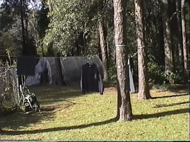 A woman pushes a dolly through a wooded backyard, followed by a young man who breakdances near a clothesline in this vintage home movie