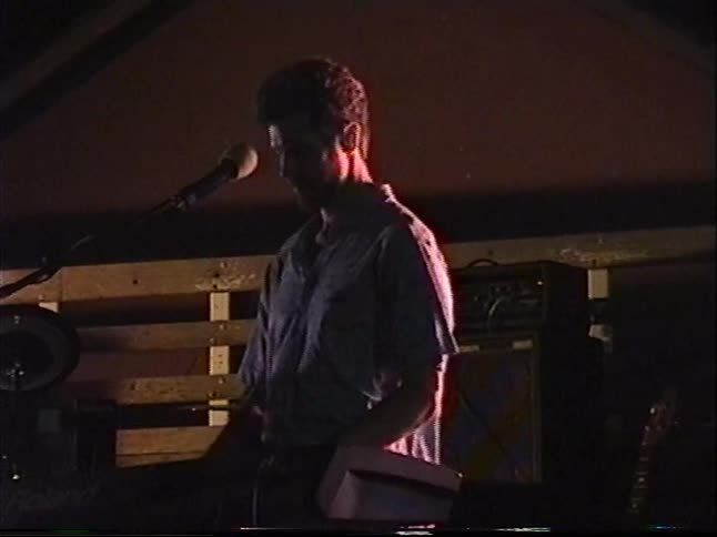 A male keyboardist sings into a microphone while performing on a dimly lit stage in the 1990s