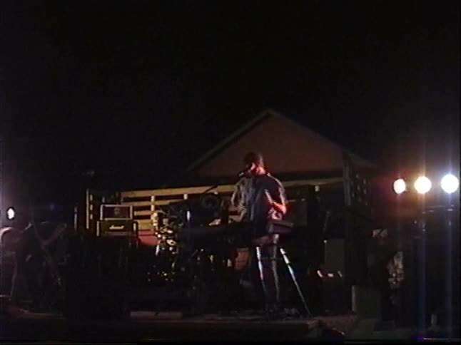 Vintage VHS footage shows a band performing on an outdoor stage at night, starting with a close-up on the keyboardist singing