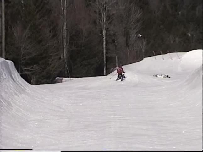 A skier in a red jacket carves down a snowy half-pipe, followed by a point-of-view shot of a snowboarder catching air