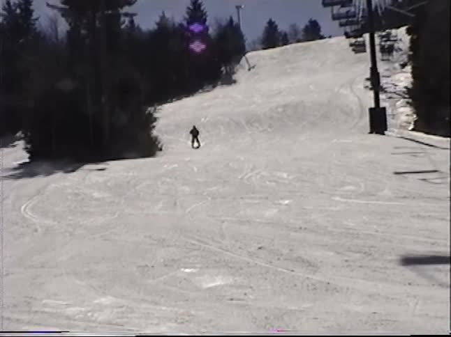 Vintage VHS footage of a person skiing down a mountain slope on a sunny day, with a ski lift in the background