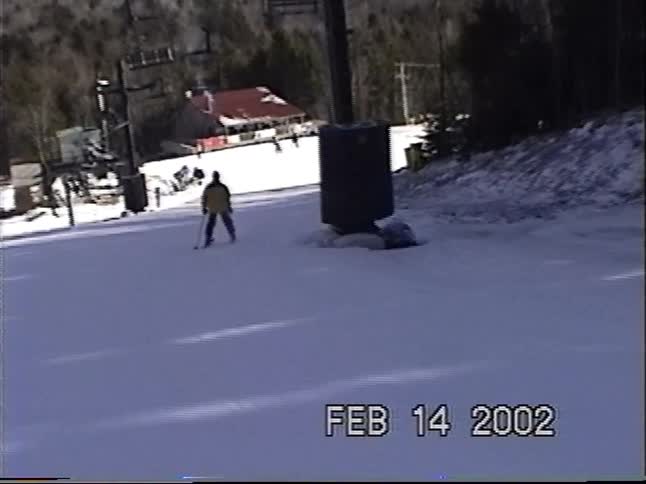 Vintage home movie footage captures a young boy skiing down a sunny, groomed mountain slope in 2002