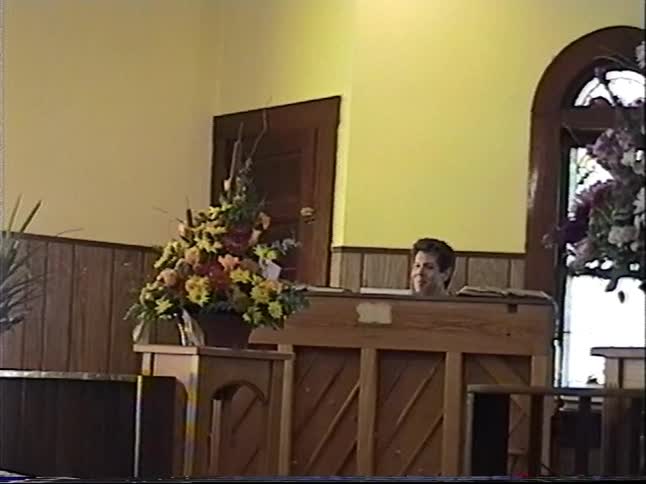 Vintage VHS footage shows a pianist in a church sanctuary, surrounded by beautiful bouquets of flowers