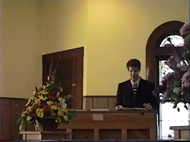 Vintage home video captures a young man in a suit reading from a book and speaking at a lectern inside a church