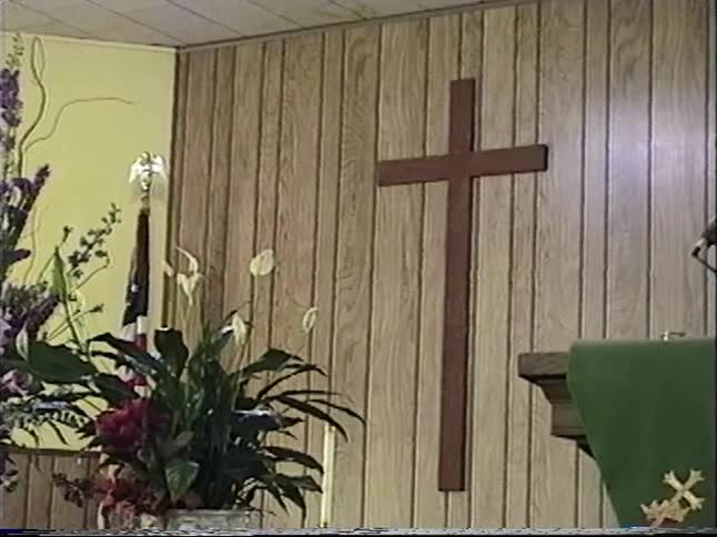A slow pan across a church altar reveals the organist, floral arrangements, and a large wooden cross on a wood-paneled wall