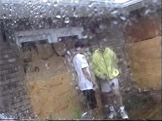 Vintage VHS footage shows two men observing a boarded-up house during a rainstorm