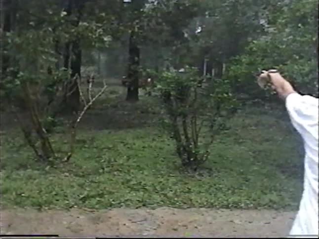Vintage home movie footage shows a young man standing in a heavy downpour and pointing a gun into the woods