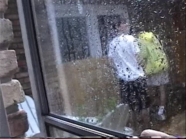 Vintage footage shows two men boarding up a house with plywood in the pouring rain as seen from inside through a wet window