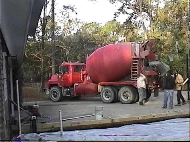 Construction workers pour a concrete foundation from a red cement mixer truck at a building site in a wooded area