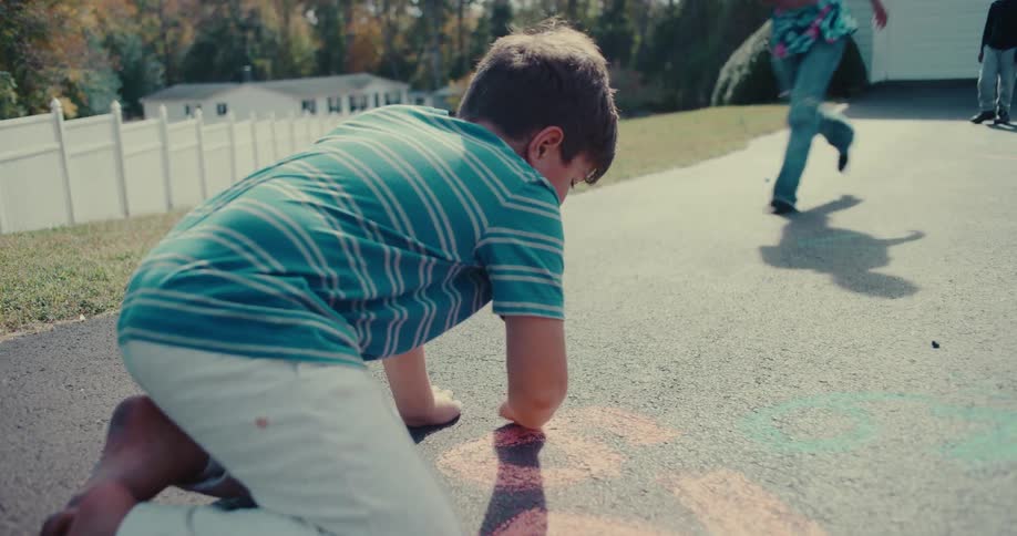 A young barefoot boy kneels on the driveway, drawing with colorful chalk in slow motion as another person plays in the background