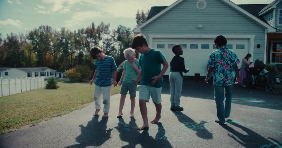 In slow motion, a diverse group of young boys enjoy a game of hopscotch in a suburban driveway on a sunny day