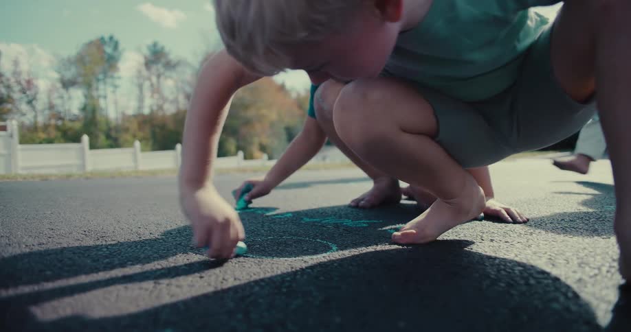 Two young boys barefoot and focused, draw together with chalk on a sunny suburban driveway