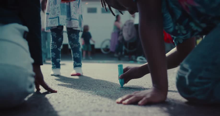 A close-up shot shows children's hands drawing with colorful chalk on the asphalt during a sunny day