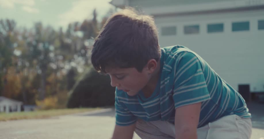 A young barefoot boy in a striped shirt concentrates while drawing with turquoise chalk on an asphalt driveway