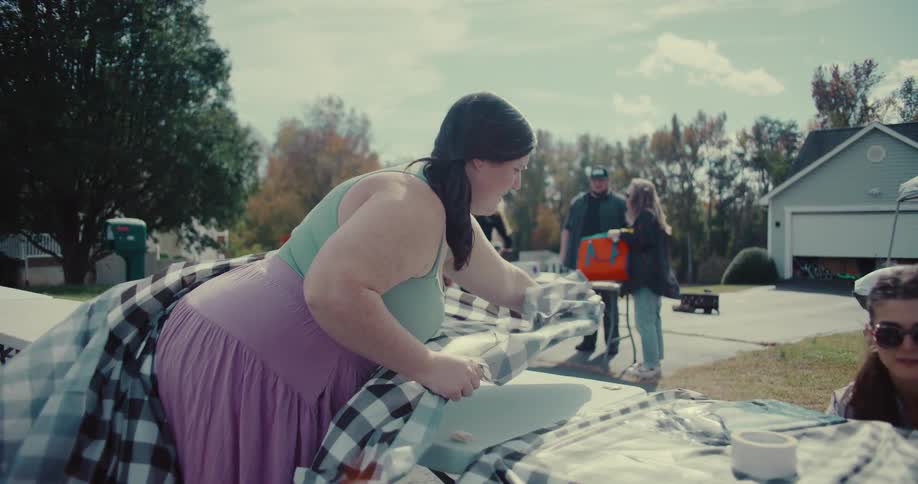 Two women lay a checkered tablecloth on a long table in slow motion as they prepare for a neighborhood party on a sunny day