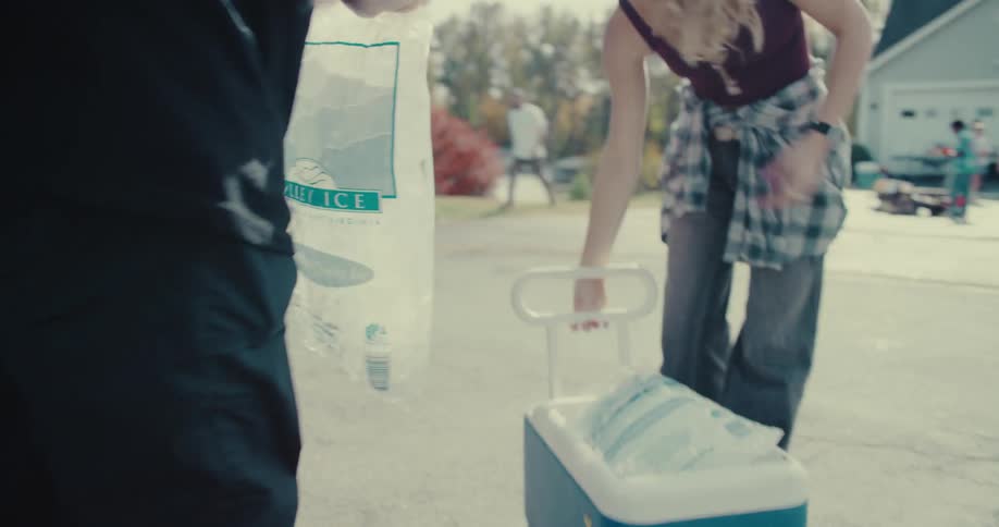 A young man and woman load bags of ice into a cooler while preparing for a neighborhood block party on a sunny day