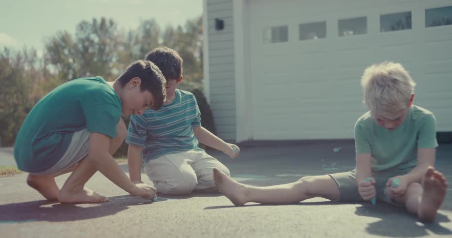 Three young boys enjoy a sunny day drawing with colorful sidewalk chalk on the driveway
