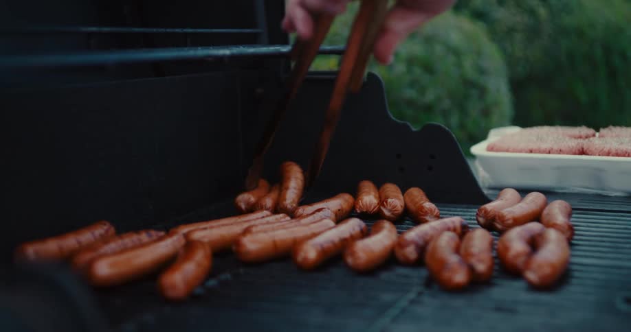 A close-up of a person using tongs to flip sausages and hot dogs on a hot barbecue grill in the backyard
