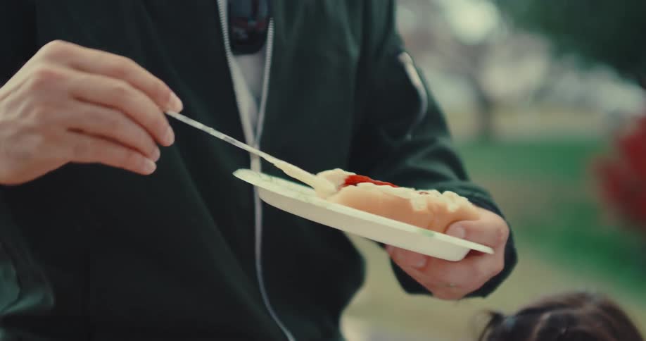 A close-up slow-motion shot captures a person carefully spreading mayonnaise onto a hot dog with ketchup at an outdoor picnic