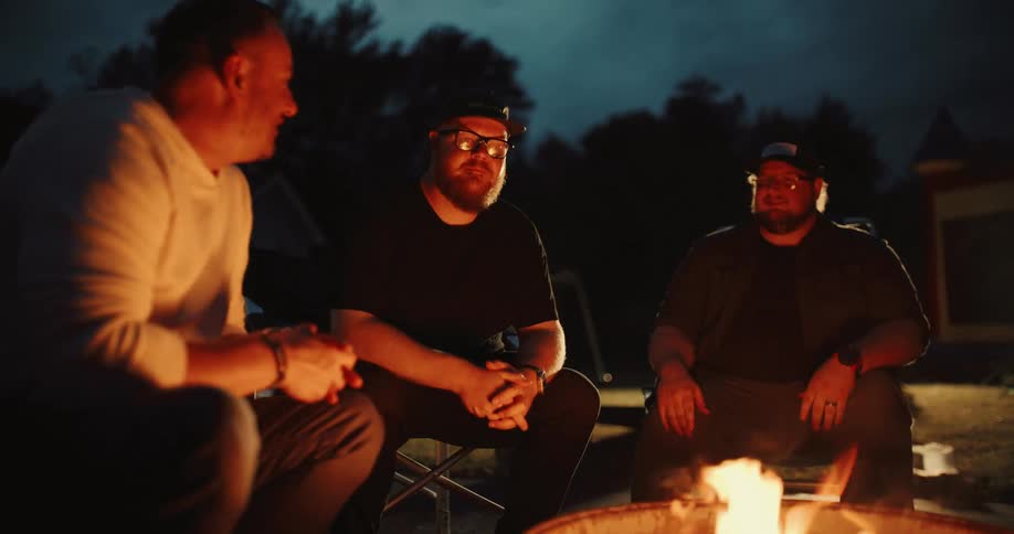 Three male friends sit together and have a deep conversation around a bonfire at night