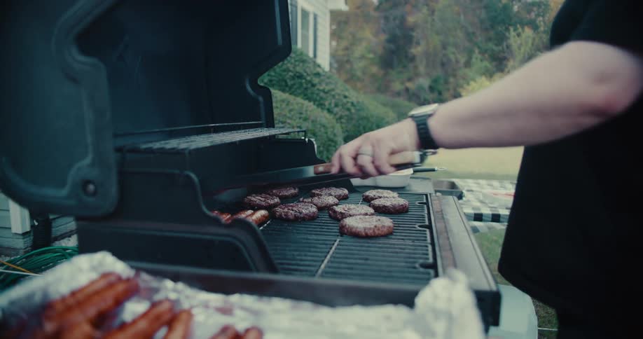 A man flips burgers and hot dogs on a smoky, flaming barbecue grill during a backyard cookout