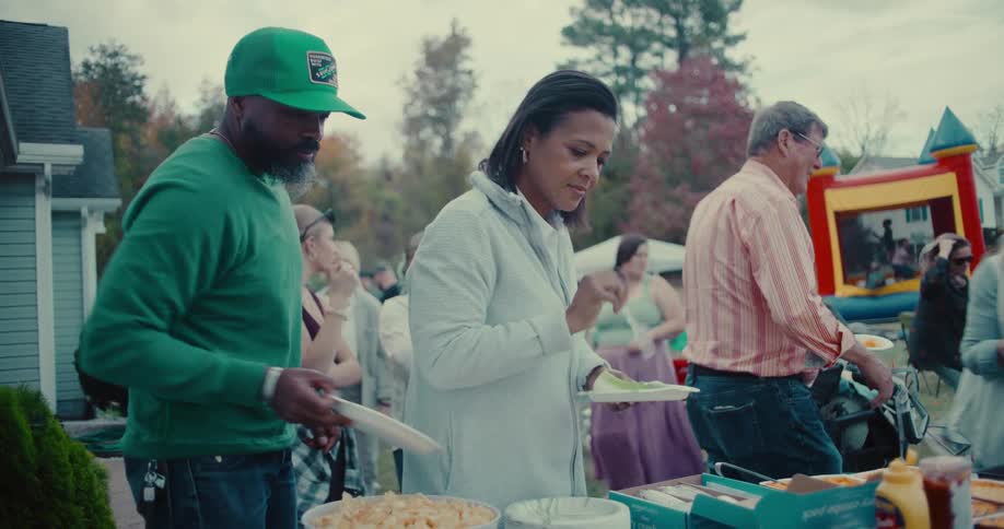 A diverse group of people get food from a potluck table at a backyard neighborhood party