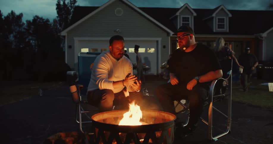 Two friends chat by a warm fire pit during an outdoor party at a suburban home in the evening