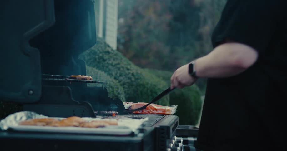 A man flips burgers on a flaming barbecue grill with a spatula during an outdoor cookout in slow motion