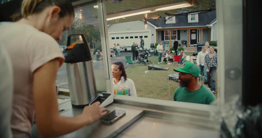 A view from inside a food truck captures diverse neighbors ordering food at a friendly suburban block party