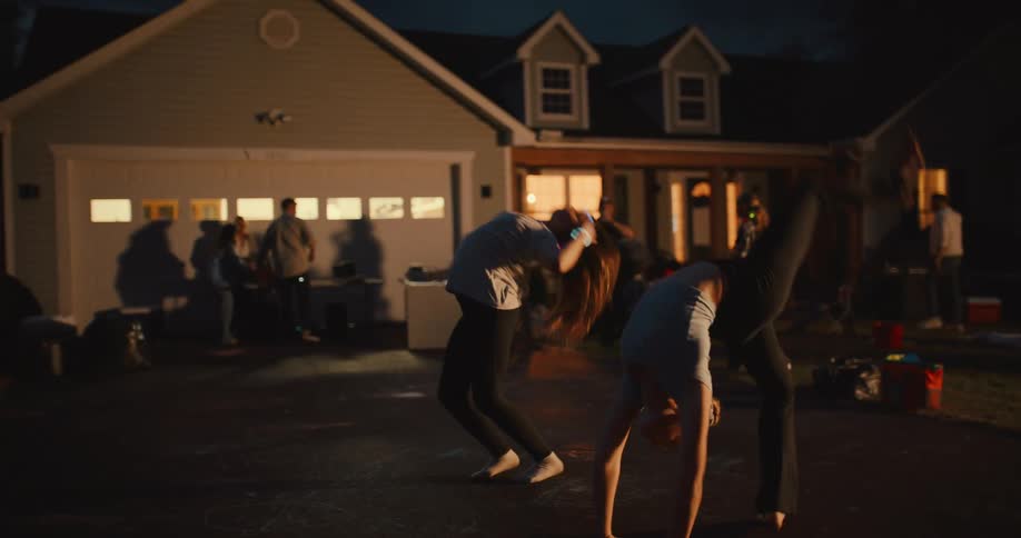In slow motion, two athletic teenage girls wearing glow sticks perform synchronized cartwheels and backbends in a driveway during a fun neighborhood block party at night