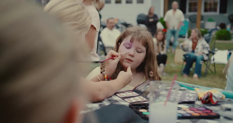 A girl gets her face painted by a friend in slow motion at an outdoor summer party with bubbles floating by